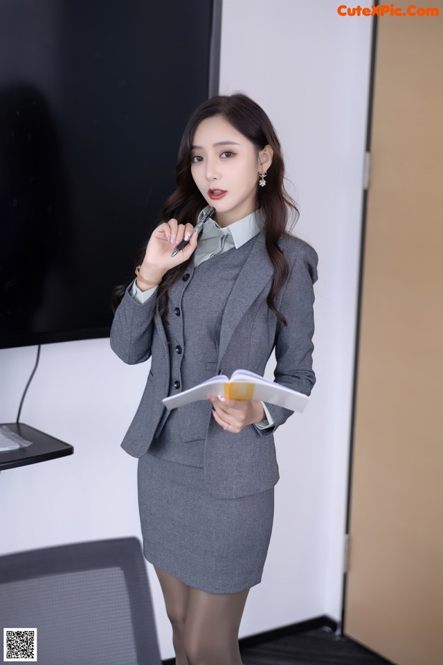 A woman in a gray suit holding a pen and a book.